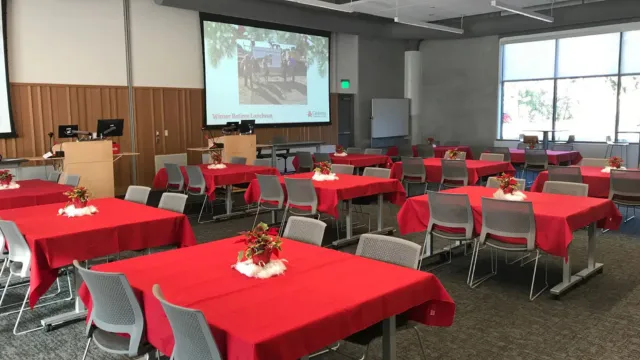 A conference room with red tablecloth-covered tables arranged in rows. Each table has a floral centerpiece. A large screen displays a presentation.