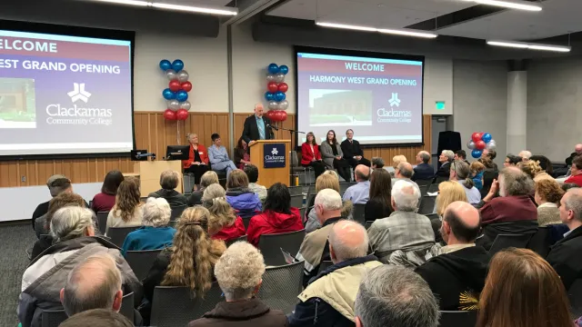 Audience at Clackamas Community College's Harmony West Grand Opening. A speaker addresses attendees, seated before a podium with two screens flanking him.