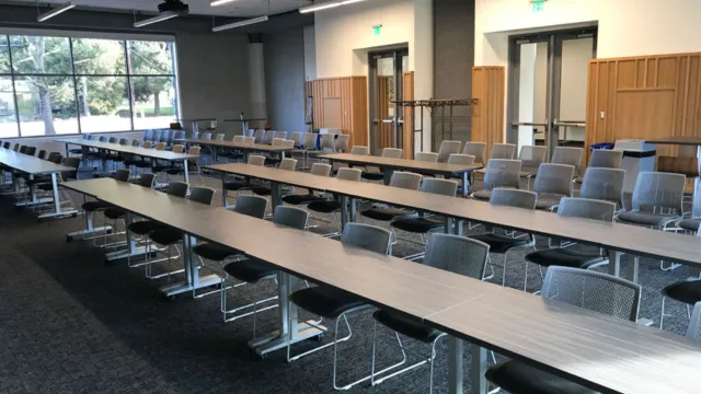 A modern, empty conference room with long tables and mesh chairs arranged in rows. Large windows let in natural light, creating a spacious atmosphere.