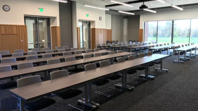 A spacious, modern conference room with long tables and rows of gray chairs, facing a large window showing a green landscape. Bright, professional ambiance.