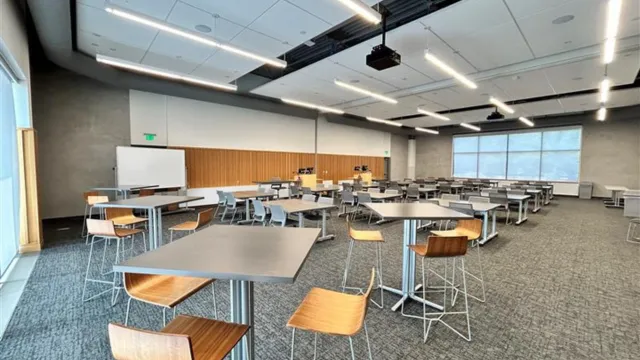 Spacious, modern conference room with gray tables, wooden chairs, ample ceiling lights, and large windows. Neutral tones create a professional atmosphere.