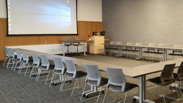 A large conference room with U-shaped tables, gray chairs, and a projector screen displaying a computer desktop. The space is modern and organized.