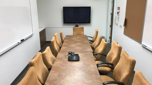 A conference room with a long table surrounded by tan chairs, a video conference device on the table, and a blank flat-screen monitor on one wall.