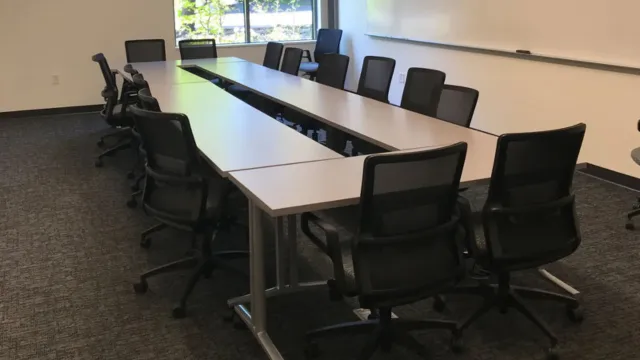 Conference room with a U-shaped table setup, surrounded by black mesh office chairs. A large window provides natural light, creating a professional atmosphere.