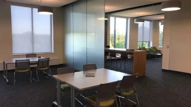 Modern study area with tables and chairs, large windows with blinds, and soft lighting. The space includes separate seating and lounge area for a calm ambiance.