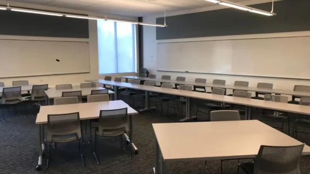 Modern classroom with rectangular tables flanked by long tables, gray chairs, whiteboards on walls, and soft natural light from a large window.