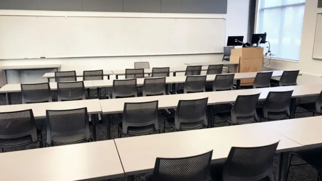 Spacious, empty classroom with gray chairs and white tables facing a large whiteboard. A wooden podium with computers is in the front right corner.