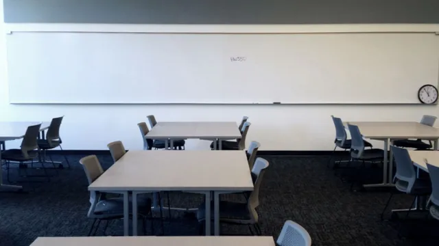 Empty classroom with several gray tables and chairs arranged in rows on a dark carpet. A large whiteboard spans the wall, with a clock to the right.