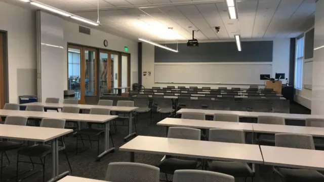 Empty classroom with rows of gray desks and chairs, facing a whiteboard and digital projector. Bright lighting, modern and organized atmosphere.