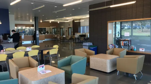A modern interior with various seating areas. Features include yellow chairs, beige and teal armchairs, tables, bookshelves, and a person at the information desk, creating a welcoming, collaborative space.