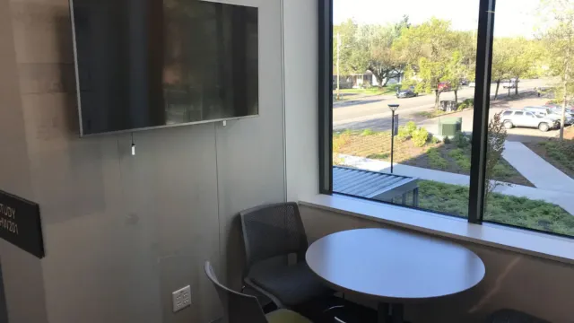 Compact office space featuring a round table, three chairs, and a wall-mounted TV. Large windows provide a sunny view of trees and a parking lot.