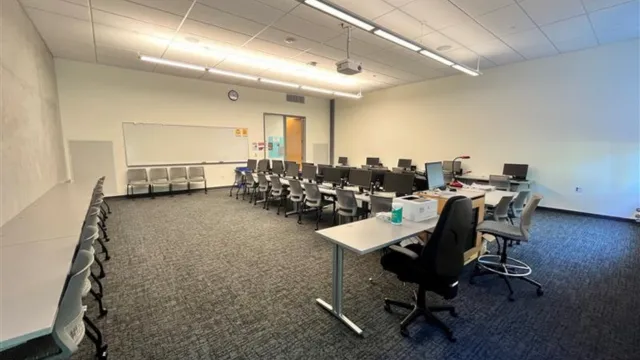 A modern classroom with rows of computers on desks, chairs aligned, whiteboard, and projector. The atmosphere is organized and well-lit.
