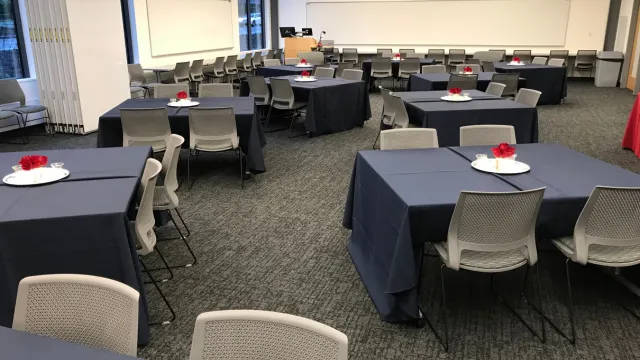 A conference room with neatly arranged tables covered in dark tablecloths, each adorned with a white plate and red centerpiece, creating a formal atmosphere.