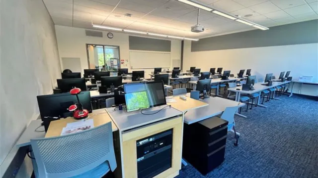 A modern computer lab with rows of desks, each featuring a computer monitor and chairs. Bright overhead lighting and a projector create a studious atmosphere.