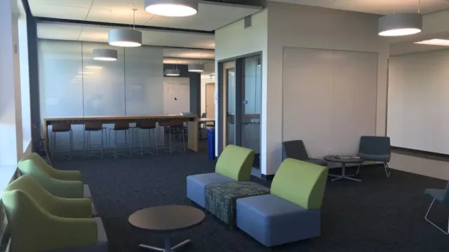 Modern office lounge with cozy seating, green and gray chairs, low tables, bar stools, and pendant lights. Bright and welcoming atmosphere.