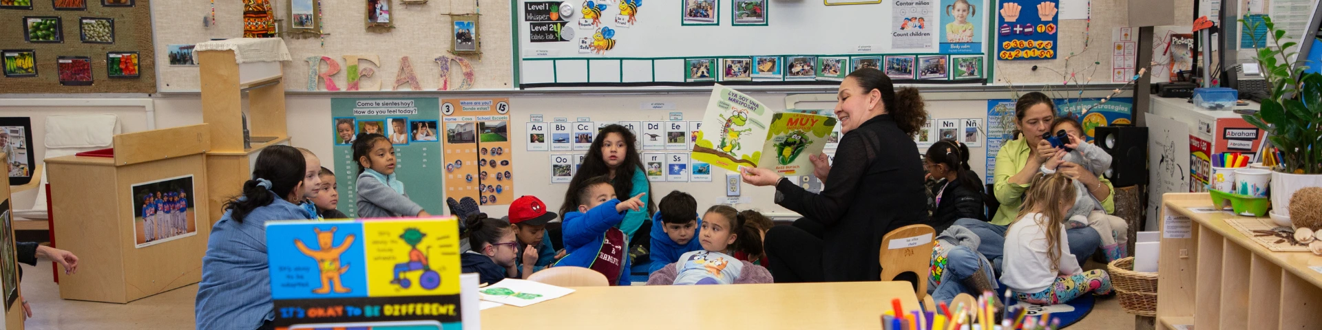 Classroom scene with children seated around a teacher reading a book aloud. "Diversity Community" is displayed prominently with colorful decor.