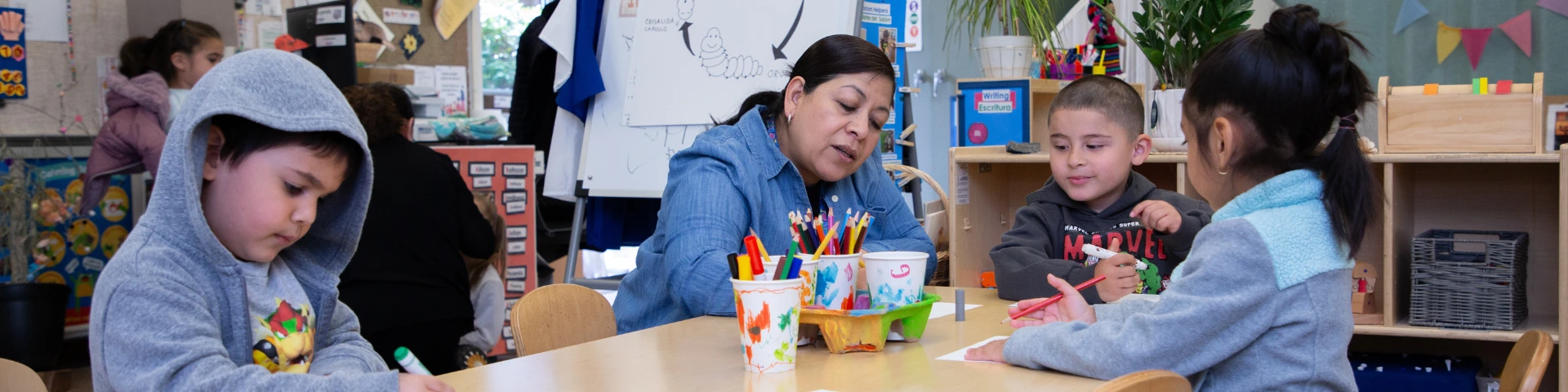 A teacher sits with three young students at a classroom table, engaging in a drawing activity. The room is colorful and decorated with plants and educational tools. The atmosphere is focused and collaborative.