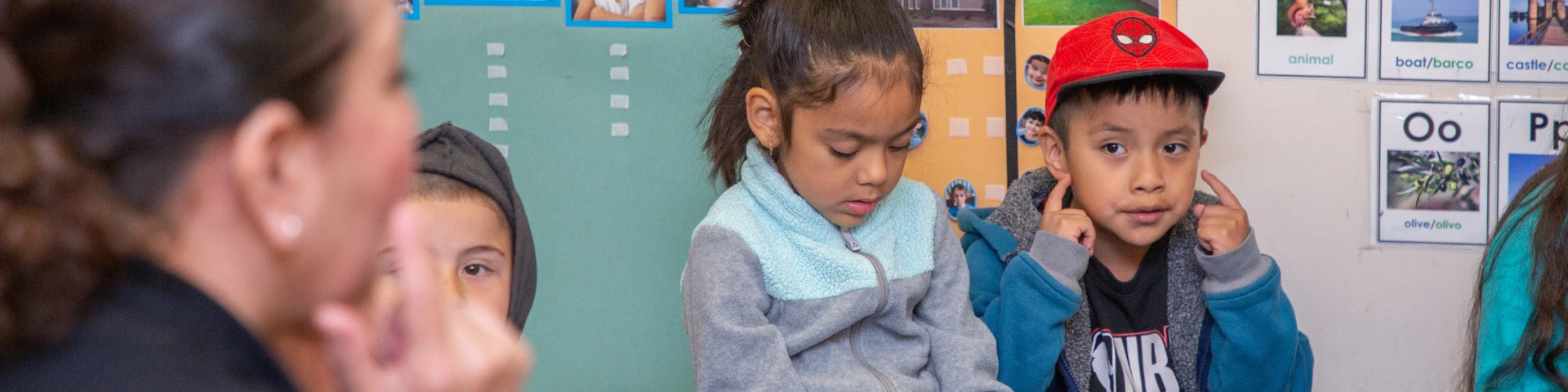 A teacher sits on the floor with three young children in a classroom. The walls display colorful educational posters.