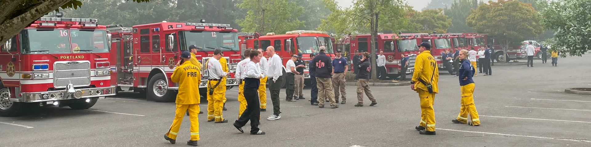 A diverse group of emergency management personnel — ranging from firefighters to EMTs to incident managers — standing in a parking lot during wildfires making emergency coordination plans.