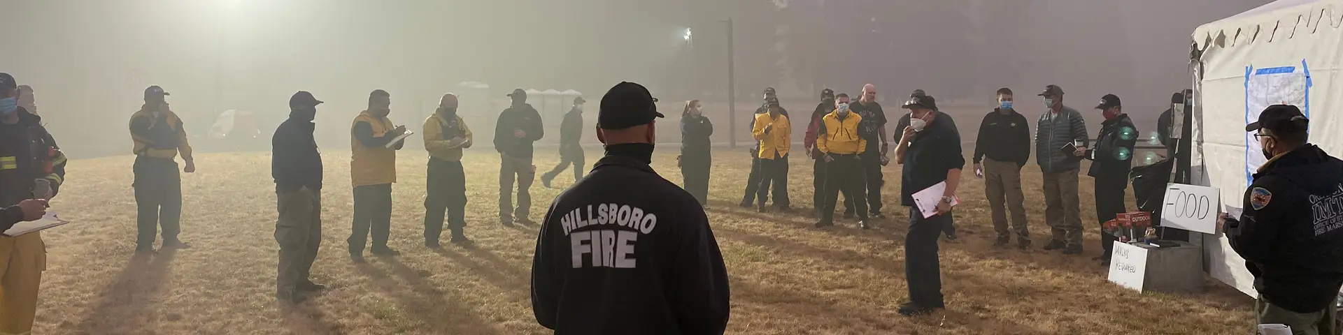 A diverse group of emergency management personnel — ranging from fire fighters to EMTs to incident managers — standing in an open field during wildfires making emergency coordination plans.