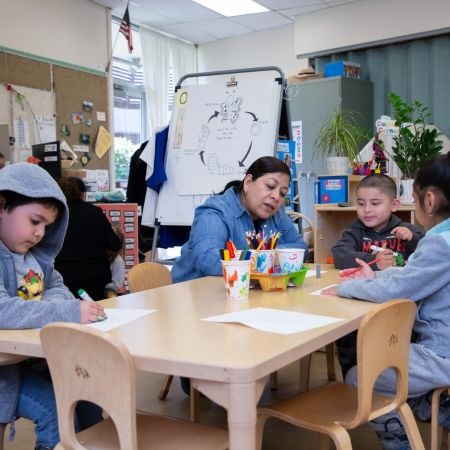 A teacher sits with three young students at a classroom table, engaging in a drawing activity. The room is colorful and decorated with plants and educational tools. The atmosphere is focused and collaborative.