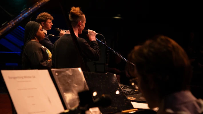 Three vocalists perform on a dark stage accompanied by a piano player in the foreground