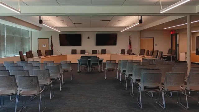 A tidy conference room is shown, featuring a U-shaped table set up with office chairs. Rows of chairs for an audience face the table, with two large screens and flags displayed at the rear, creating a professional setting.