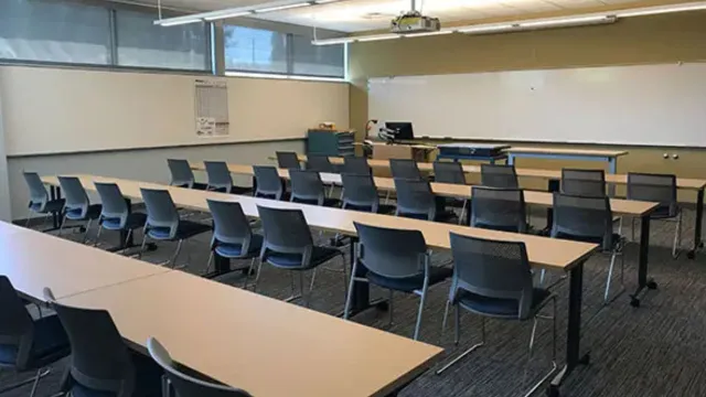 Empty classroom with rows of gray chairs and light wood tables facing a whiteboard. A projector hangs from the ceiling, creating a quiet, academic atmosphere.