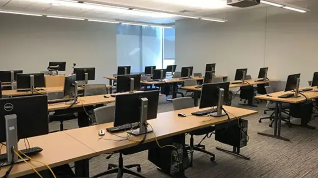 Spacious computer lab with rows of empty desks, each equipped with computers and chairs. Soft lighting and a large window create a calm, orderly atmosphere.