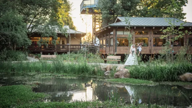 A bride and groom stand by a tranquil pond, embraced in a lush garden. Wooden cabins are in the background, set against a setting sun, creating a romantic and serene atmosphere.