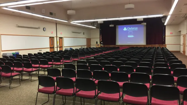 Empty conference room with rows of black and red chairs facing a large screen displaying a logo. Bright lighting and a neutral tone create a calm atmosphere.
