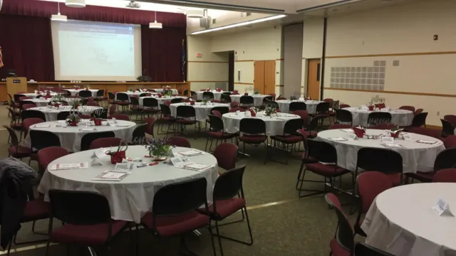 A large conference room set up for an event with round tables covered in white cloths, red chairs, scattered programs, and floral centerpieces, exuding an elegant atmosphere.