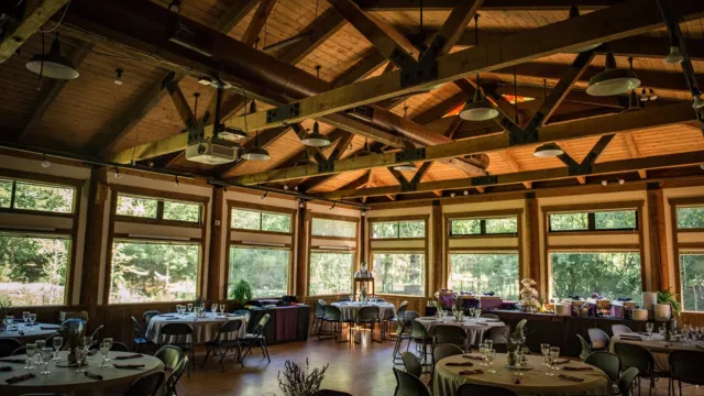 Rustic wooden hall with large windows and vaulted beams, set for an event. Round tables with chairs, tableware, and soft daylight create a welcoming ambiance.