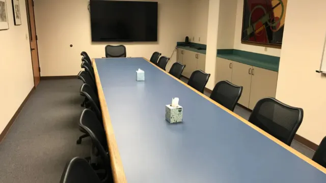 A long conference table with black chairs in a meeting room, featuring a wall-mounted TV, abstract art, tissue boxes, and cabinets, creating a professional atmosphere.
