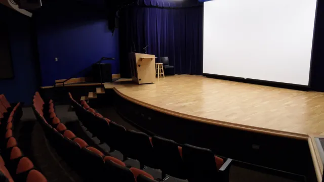 An empty auditorium features red-cushioned seats, a wooden stage with a podium, stool, and microphone, and a large white projection screen, conveying readiness and anticipation.