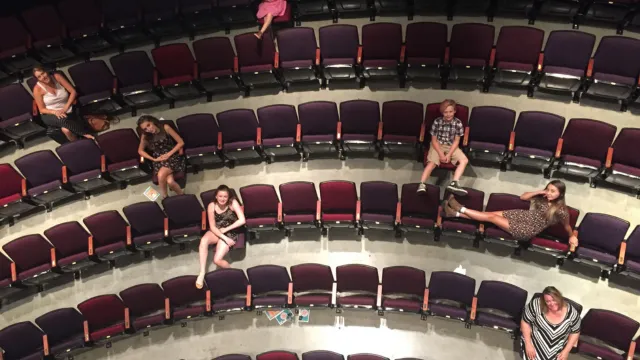 Seven people are sitting apart in a theater with rows of red and purple seats, creating an isolated, contemplative mood.