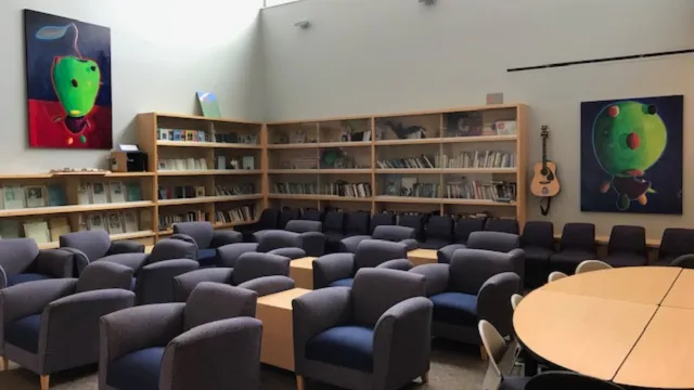 A library with gray armchairs arranged around light wooden tables. Bookshelves full of books line the wall, with colorful abstract paintings and a guitar hanging nearby.