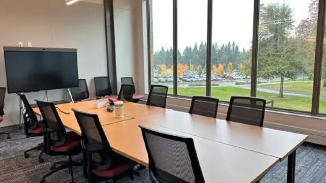 A conference room with a long table and multiple chairs faces large windows. Outside, autumn trees and a parking lot are visible, creating a calm atmosphere.
