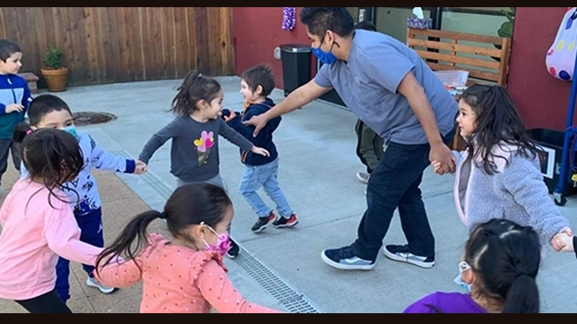 Young children hold hands and dance in a circle with their child care provider