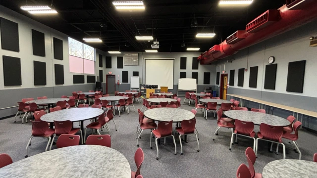 A spacious meeting room with round tables surrounded by red chairs. A large screen is at the front, beneath bright overhead lights and red ductwork.