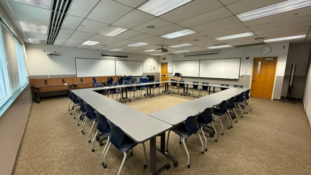 A conference room with long tables and blue chairs arranged neatly. Whiteboards and a projector screen adorn the walls under bright ceiling lights.