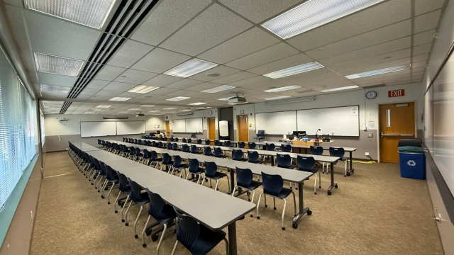 A large, empty classroom with rows of long tables and blue chairs facing whiteboards. Overhead lighting and windows illuminate the neutral-toned space.