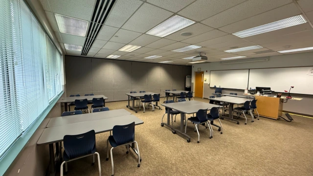 A spacious classroom with rows of grey tables and blue chairs, bright fluorescent lighting, a large whiteboard, projector, and windows with blinds.