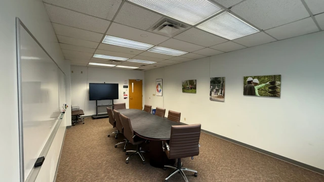 A conference room with a long oval table, surrounded by brown chairs. A large TV is mounted on the left wall, and nature photos hang on the right wall.