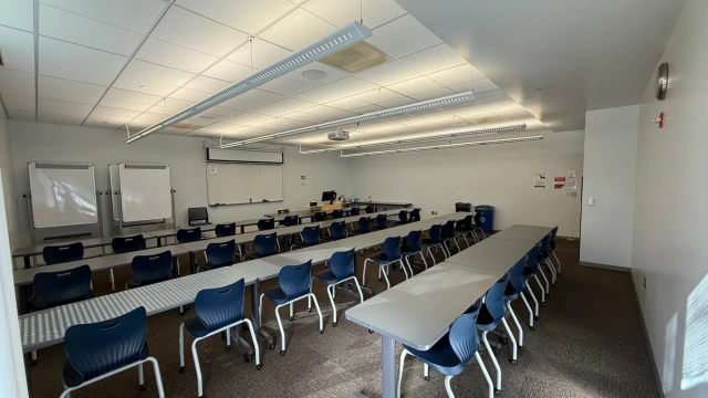Spacious classroom with rows of blue chairs and long desks, whiteboards on the front wall, a ceiling-mounted projector, and bright fluorescent lighting.