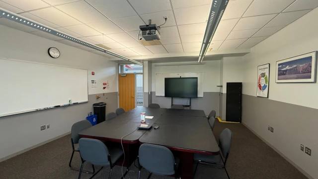 Meeting room with a large rectangular table, surrounded by chairs. Features a wall-mounted TV, whiteboard, clock, and overhead projector. Calm and organized setting.