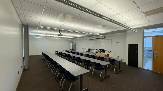 A spacious, well-lit classroom with long rows of blue chairs and tables facing a lecturer’s desk with computer equipment, and a large whiteboard.