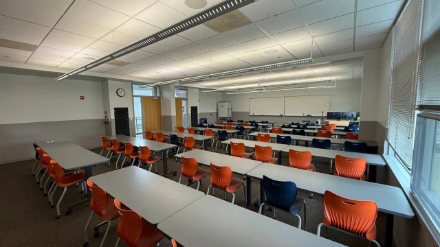 A spacious classroom with rows of white desks and alternating orange and blue chairs. Large windows on the right allow natural light, creating a bright, inviting atmosphere.